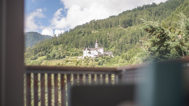 A view of a white building with a tower, surrounded by lush green hills and trees. - Lapis Monti - Apartments & Suites  (Malles Venosta)