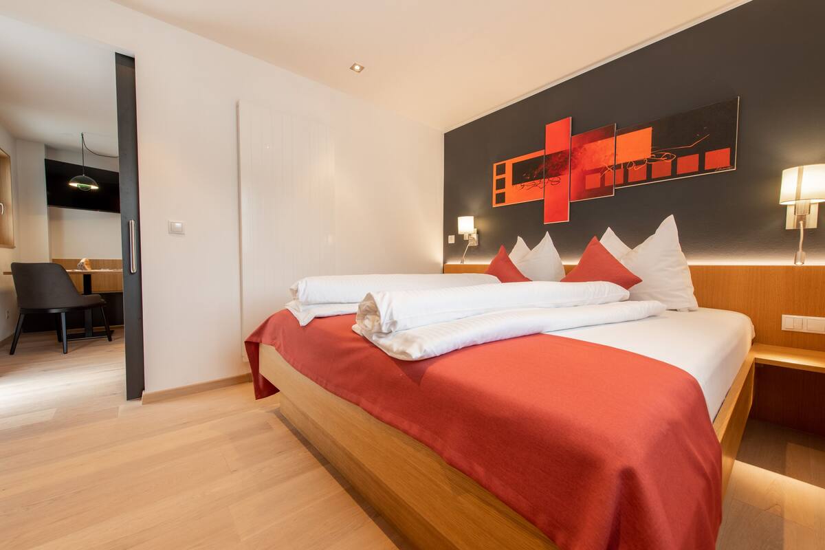 A hotel room with a wooden bed, red bedding, a modern wall art piece, and a desk with a chair.