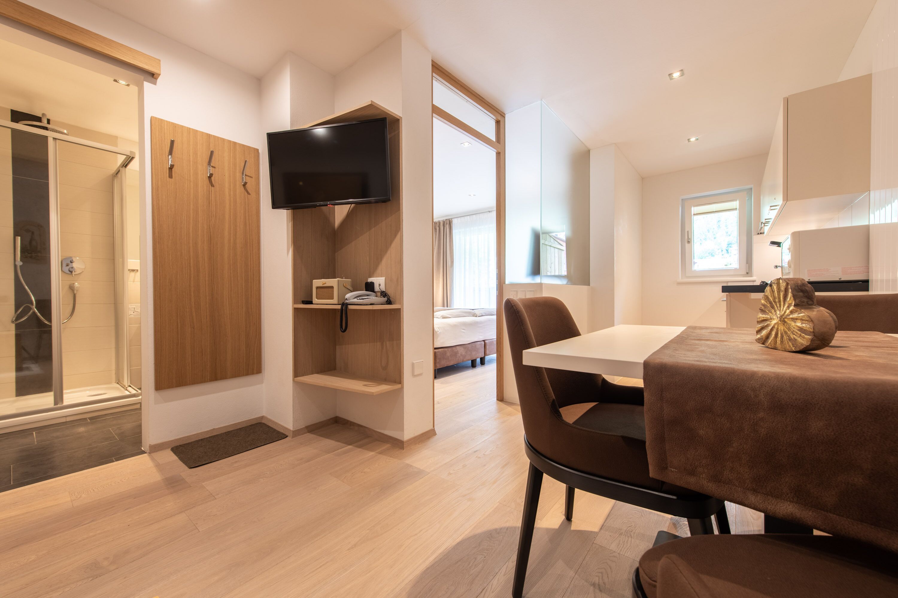 Et moderne hotelværelse med bruser, fjernsyn monteret på væggen, spiseplads med bord og stole, samt en seng i baggrunden.