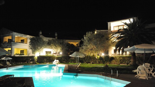 A nighttime swimming pool with lounge chairs and umbrellas, surrounded by well-lit buildings and greenery. - Niki Hotel Apartments (Rhodes)