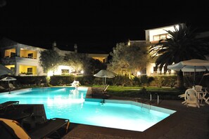 A nighttime swimming pool with lounge chairs and umbrellas, surrounded by well-lit buildings and greenery. - Niki Hotel Apartments (Rhodes)