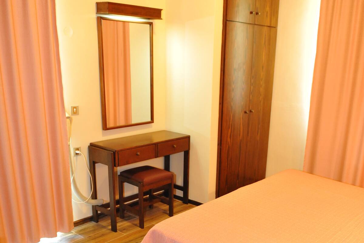 Ein Schlafzimmer mit Holzboden, einem Bett mit rosa Bettdecke, einem Holztisch mit Stuhl, einem Spiegel und einem Kleiderschrank.
