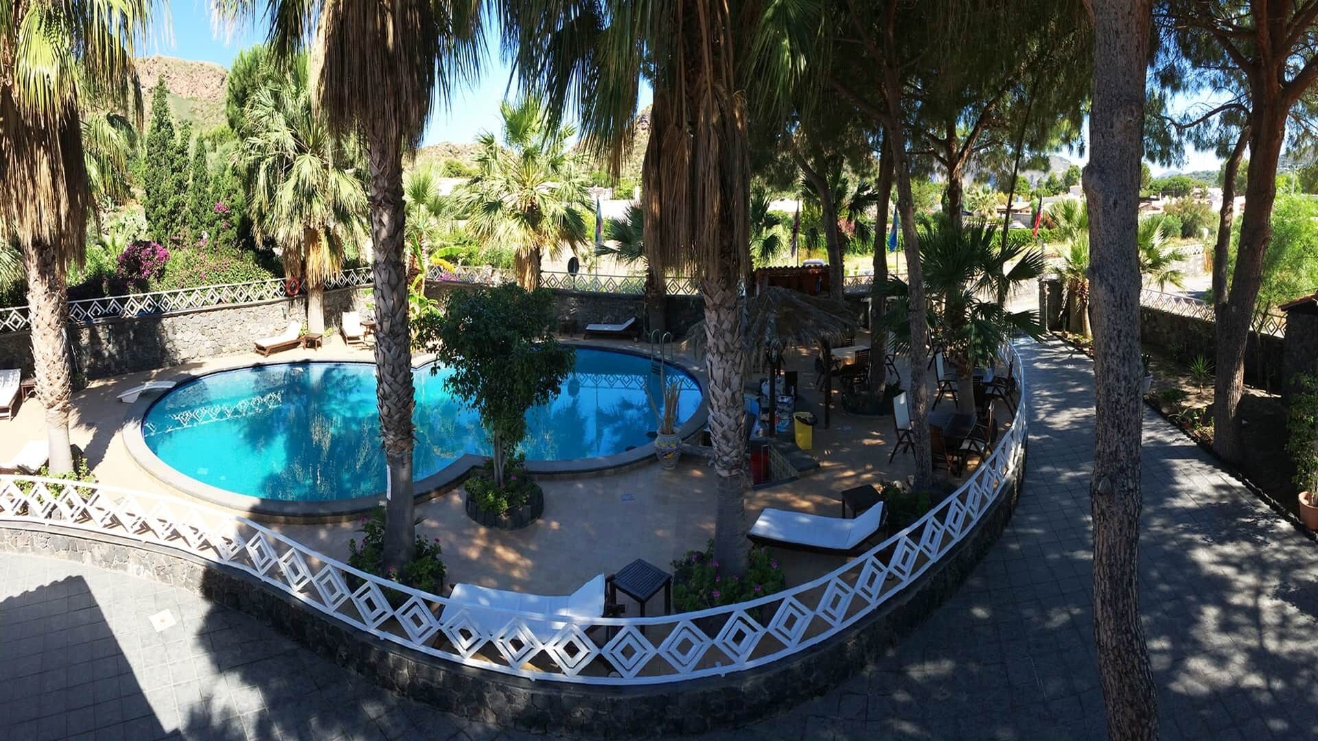 Un área de piscina con una piscina circular, rodeada de palmeras y sillas de descanso.