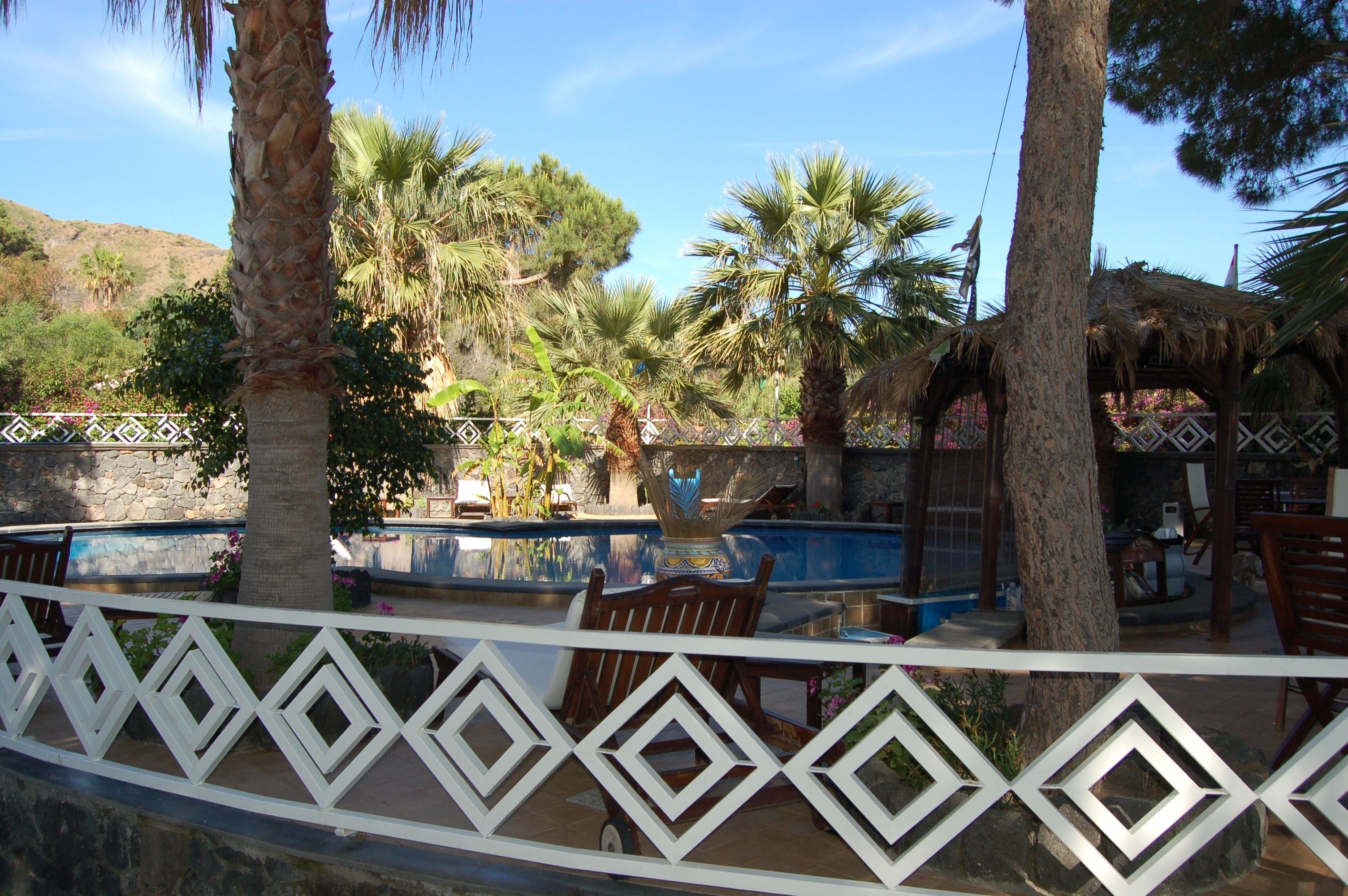 Un área de piscina con una fuente decorativa, rodeada de palmeras y una estructura con techo de paja.