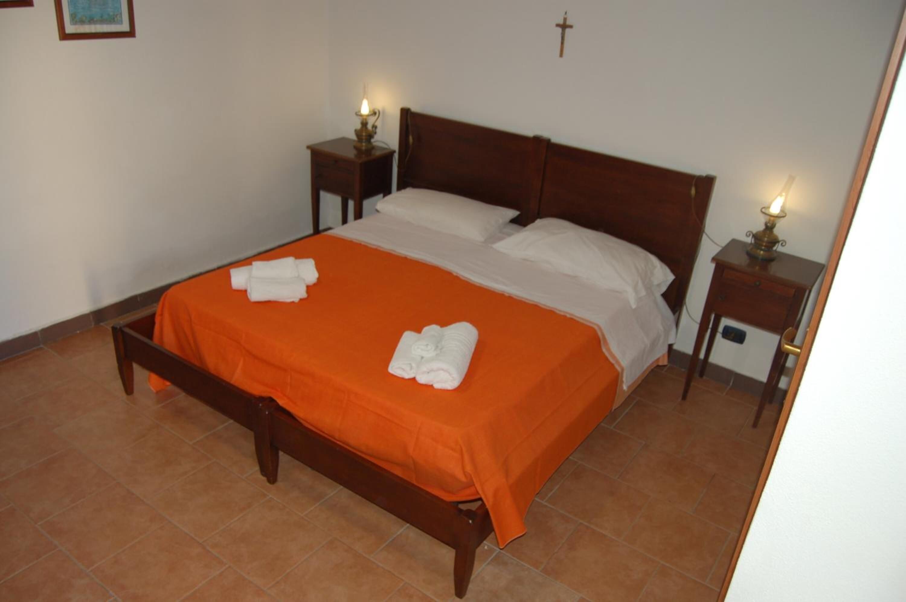 A neatly made bed with an orange bedspread and white pillows, two bedside tables with lamps, and a cross on the wall.
