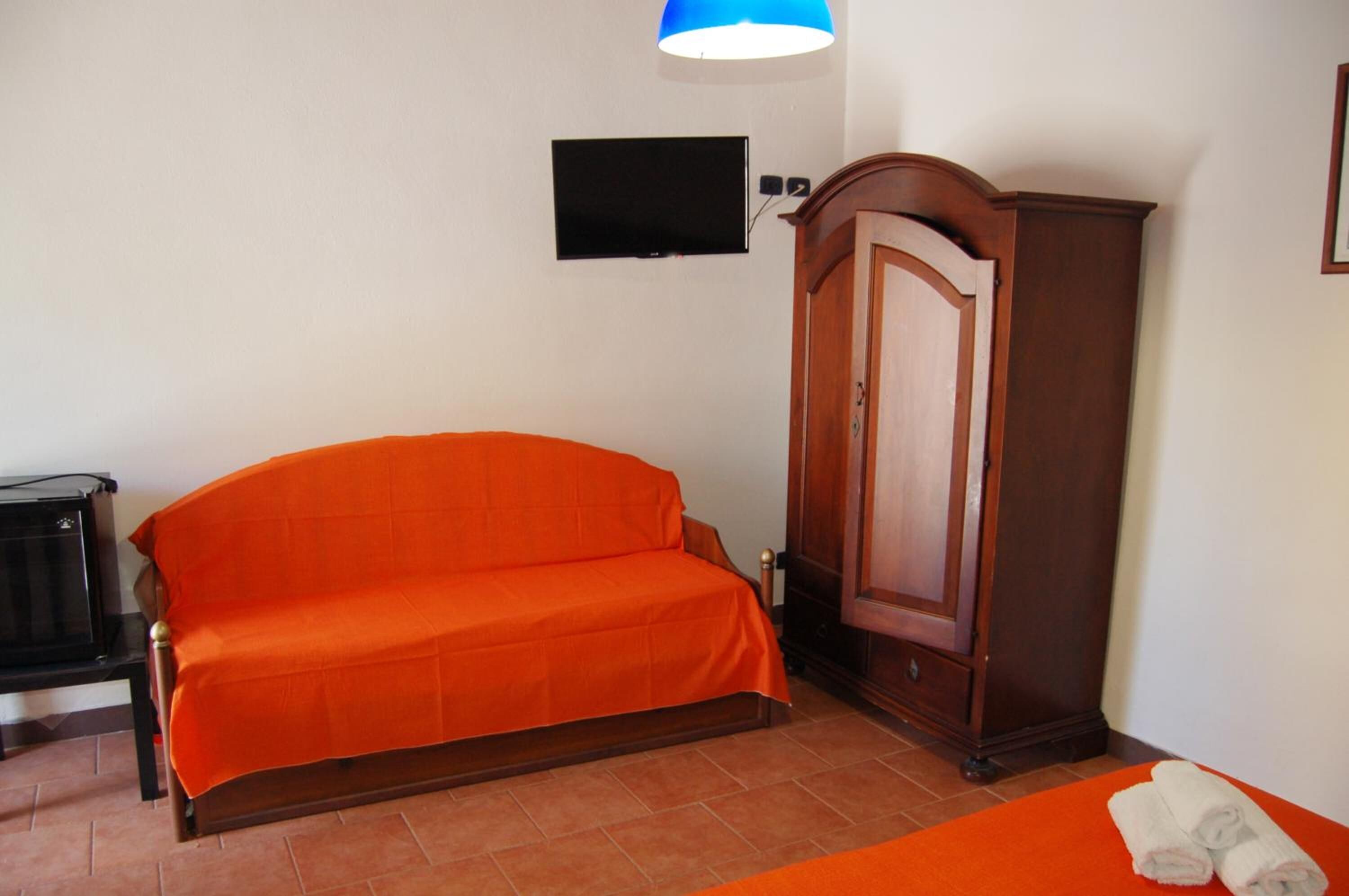 A room with a wooden wardrobe, a television mounted on the wall, and a bed with an orange bedspread.