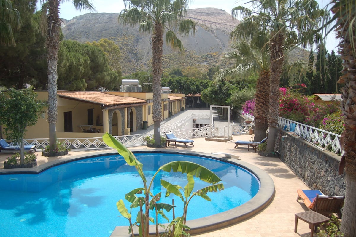A pool area with lounge chairs, palm trees, and a mountain in the background.
