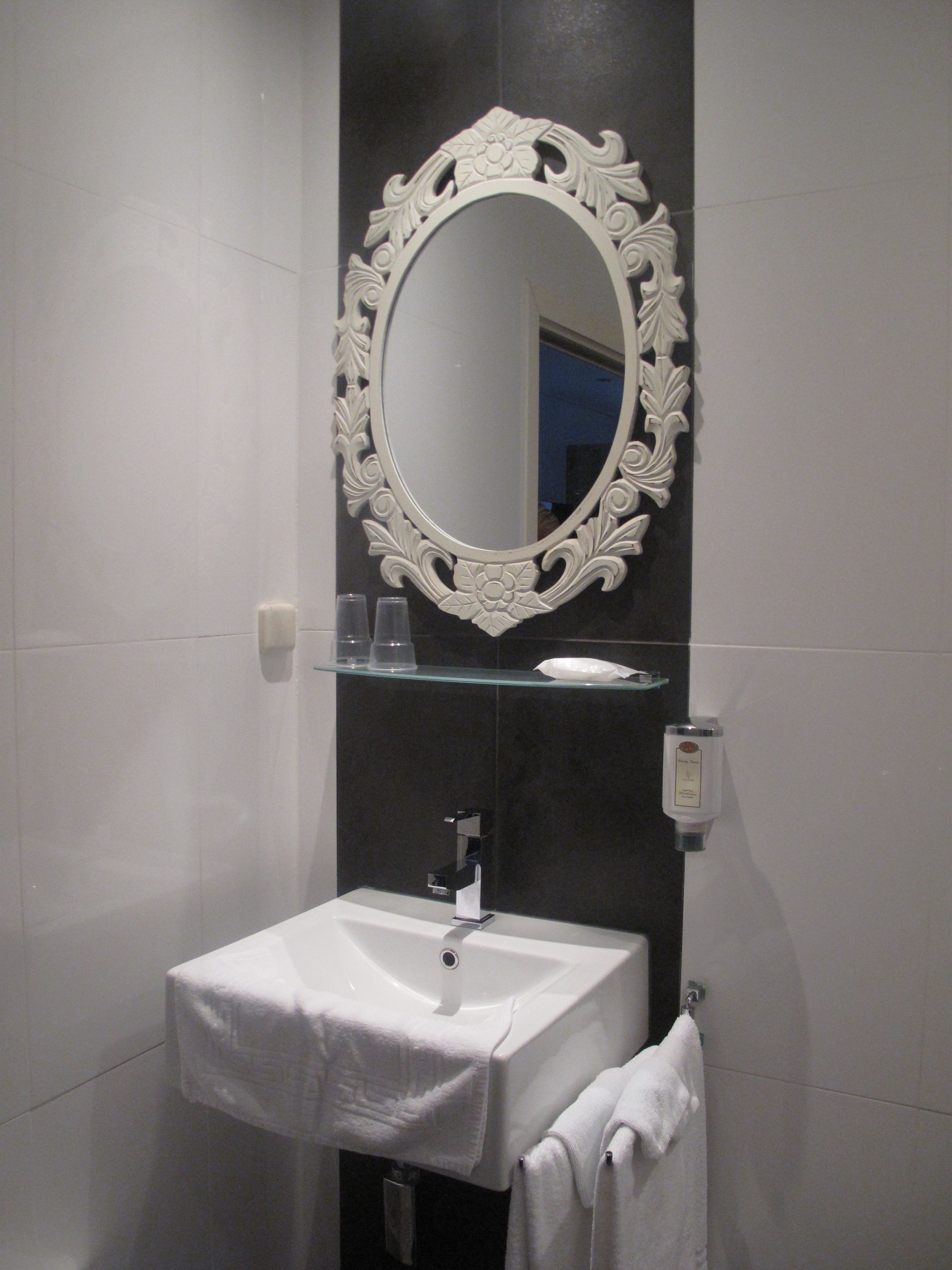 Une salle de bain avec un lavabo blanc, un miroir doté d’un cadre orné et une étagère en verre au-dessus du lavabo.