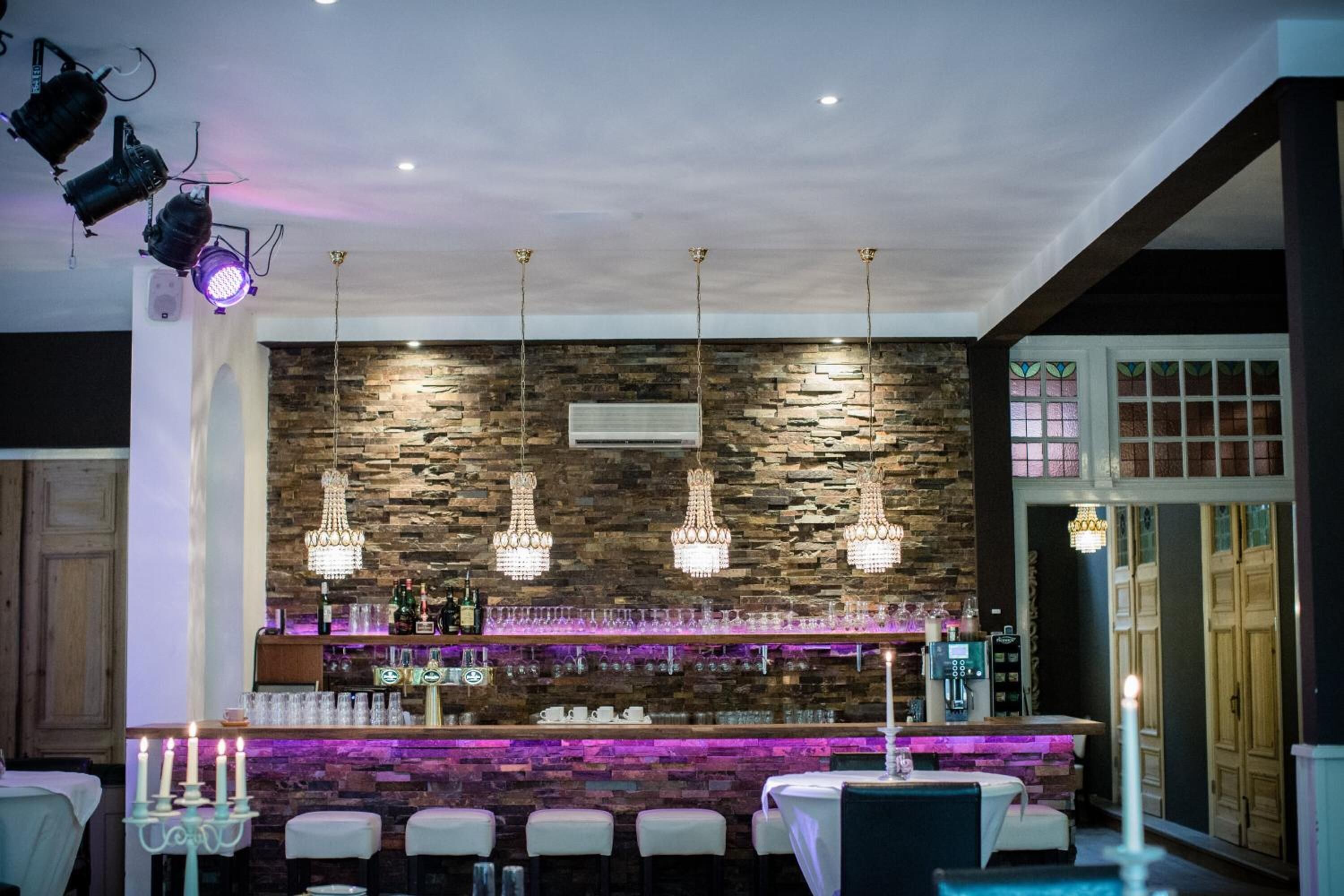 Un área de bar con un fondo de pared de piedra, varios candelabros colgantes y una barra con distintos botellas y vasos.