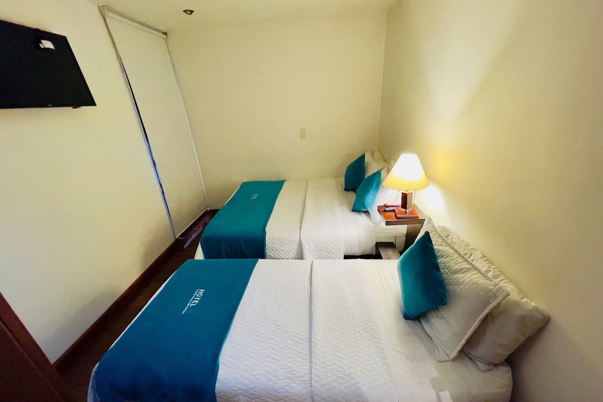 A hotel room with two beds, each with a teal bedspread and white bedding. There is a nightstand with a lamp and a remote control.