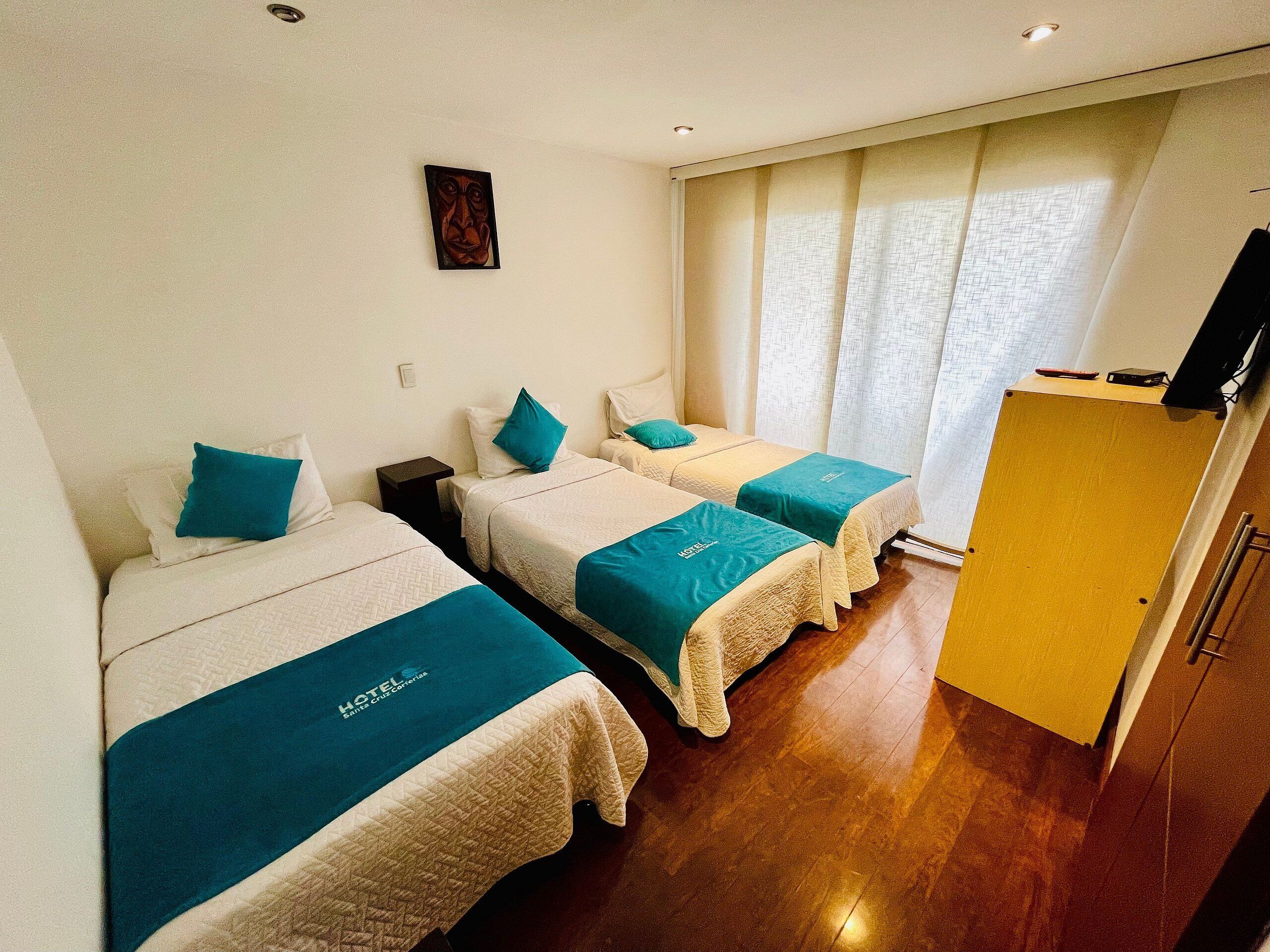 A hotel room with two beds, each with a teal bedspread and white headboard, a small table, a wall-mounted TV, and a window with curtains.