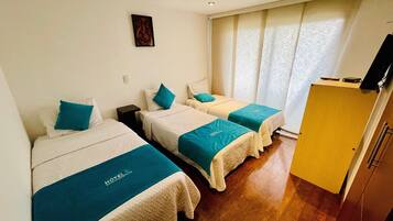 A hotel room with two beds, each with a teal bedspread and white headboard, a small table, a wall-mounted TV, and a window with curtains.