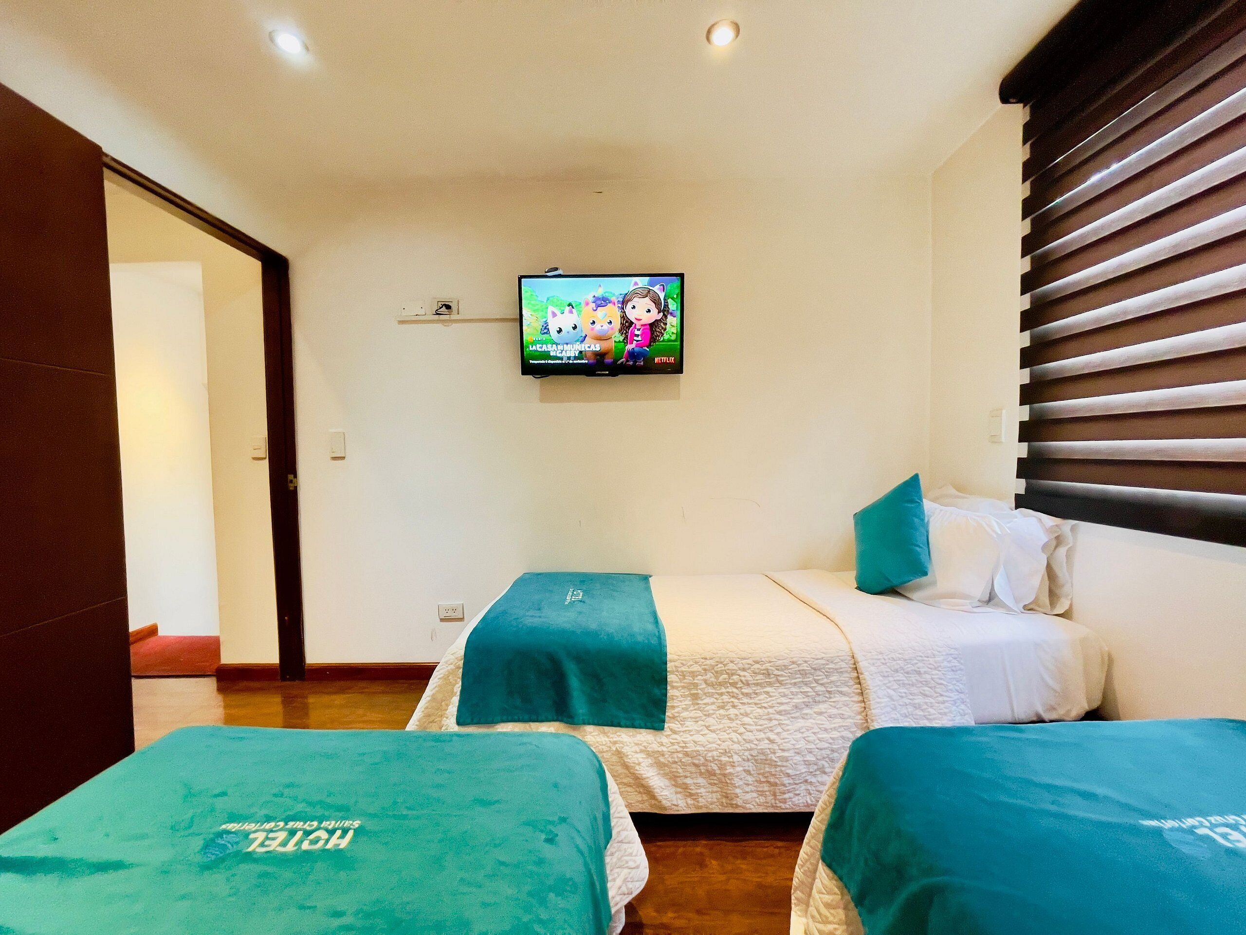 A hotel room with two beds, a TV mounted on the wall, and a window with blinds.