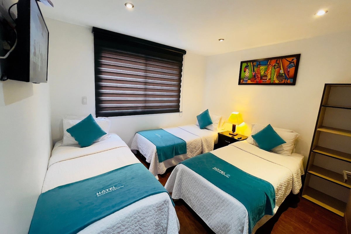 A hotel room with two beds, each with a teal bedspread and white bedding. There is a painting on the wall, a window with blinds, and a nightstand with a lamp.