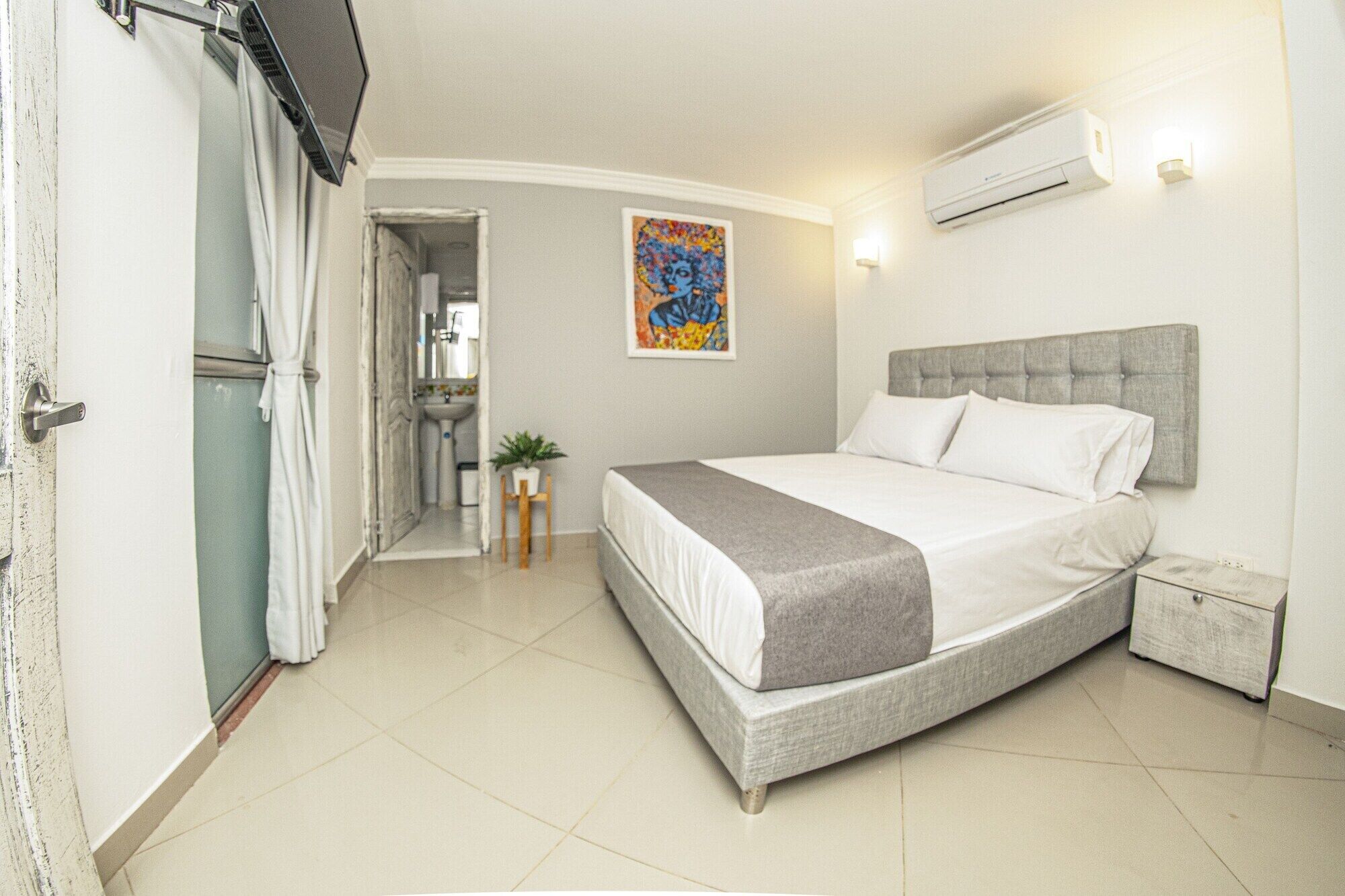 A bedroom with a bed, a nightstand, a potted plant, and a bathroom visible through an open door.
