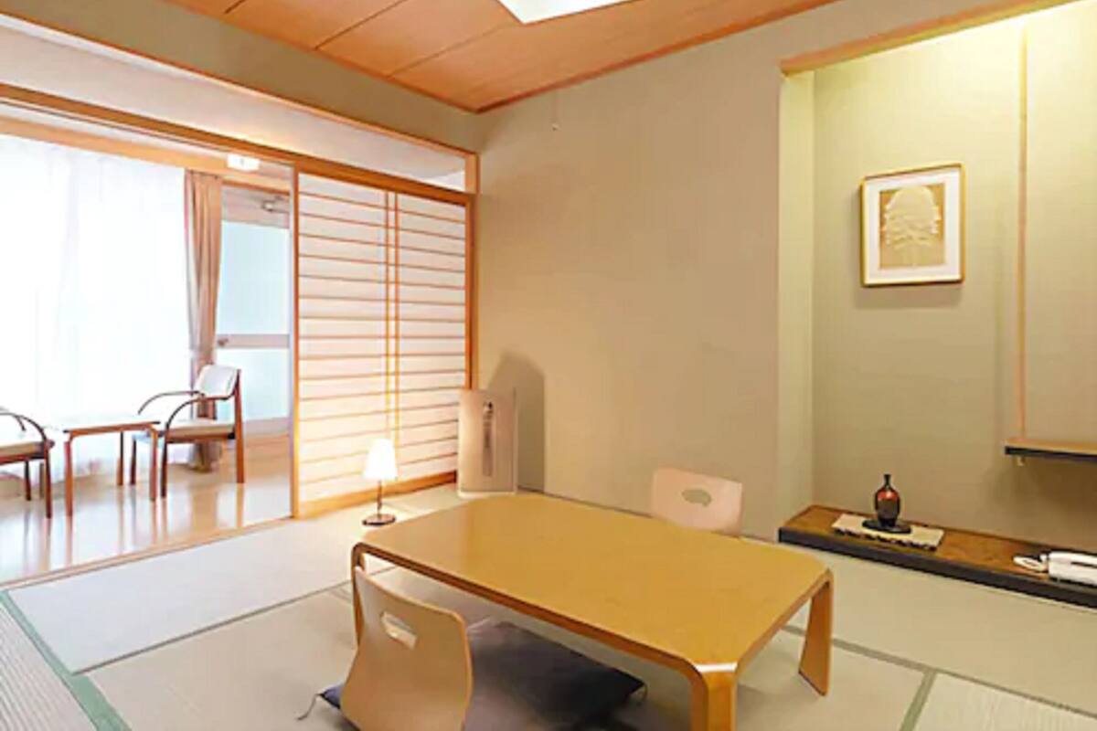 Une pièce de style japonais traditionnel, avec un sol recouvert de tatamis, une petite table en bois, une chaise, un paravent coulissant de type shoji et un petit autel orné d’un tableau encadré.