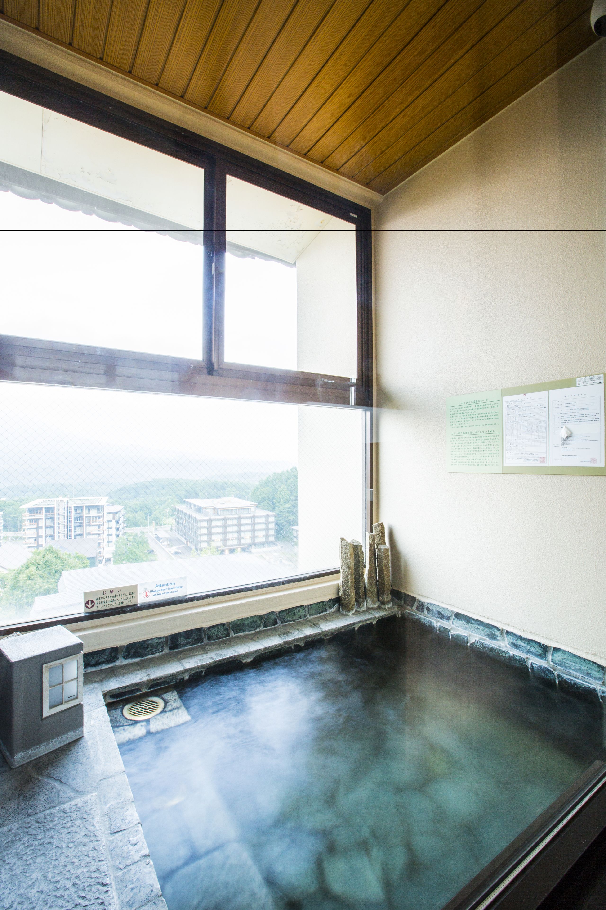 A hot spring room with a wooden ceiling, a stone floor, and a view of buildings outside.