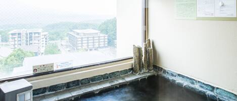 A hot spring room with a wooden ceiling, a stone floor, and a view of buildings outside.
