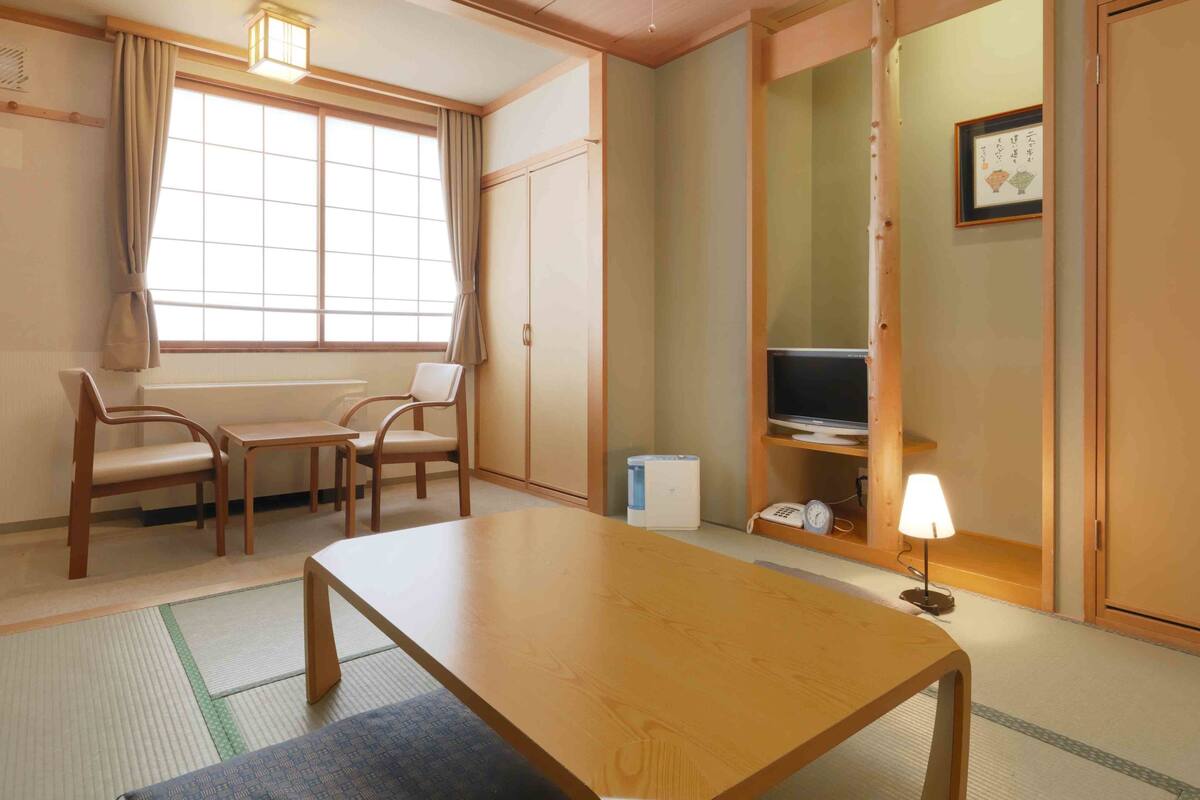 Une pièce de style japonais traditionnel, avec un sol recouvert de tatamis, une table basse, deux chaises, un téléviseur posé sur une étagère et des portes coulissantes.