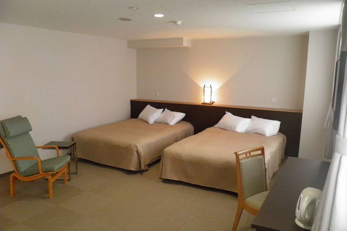 Une chambre d’hôtel avec deux lits simples, une chaise, une petite table et une table de chevet avec une lampe.