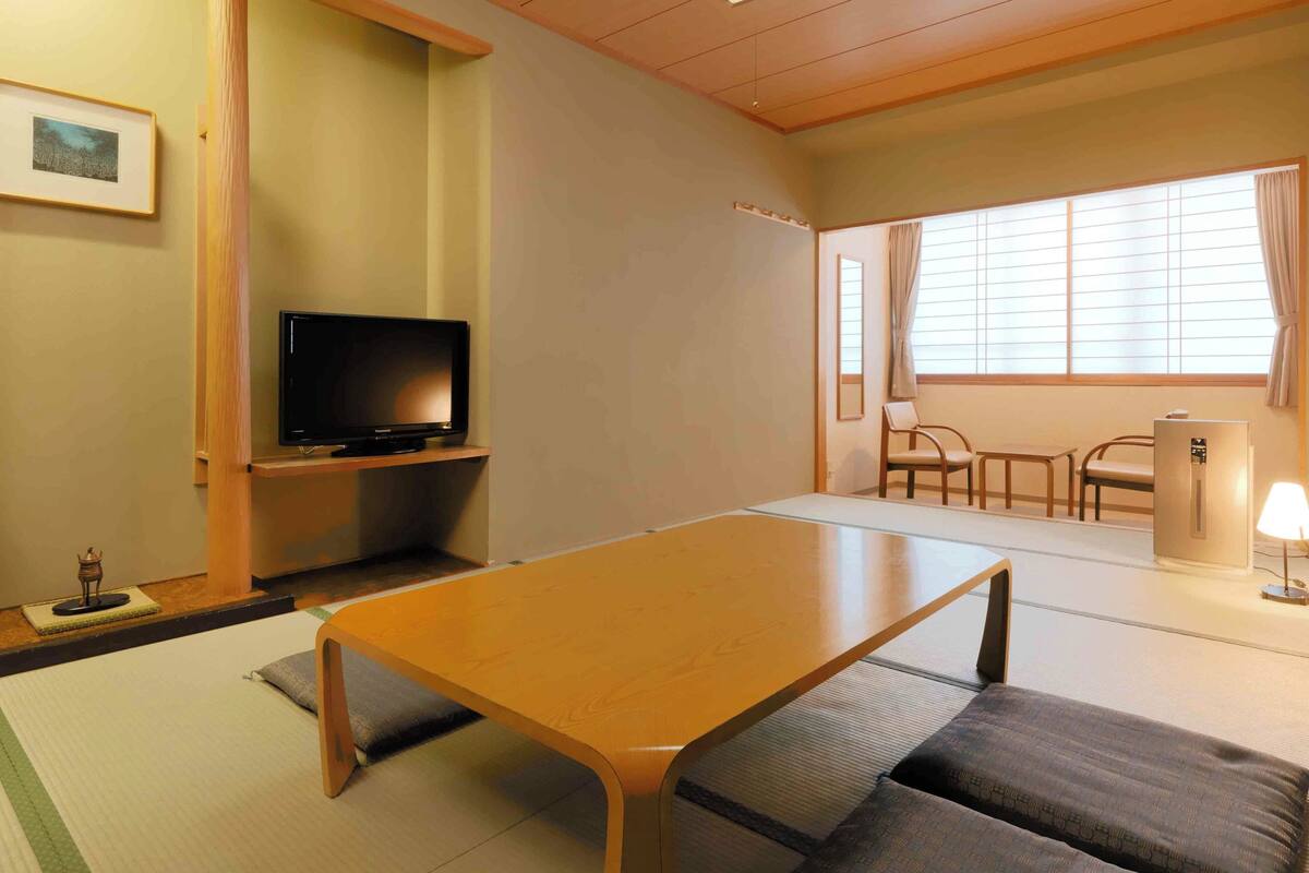 Une pièce avec une table en bois, un téléviseur posé sur un meuble, des tatamis, un canapé, une chaise, une fenêtre avec des rideaux et un petit autel.