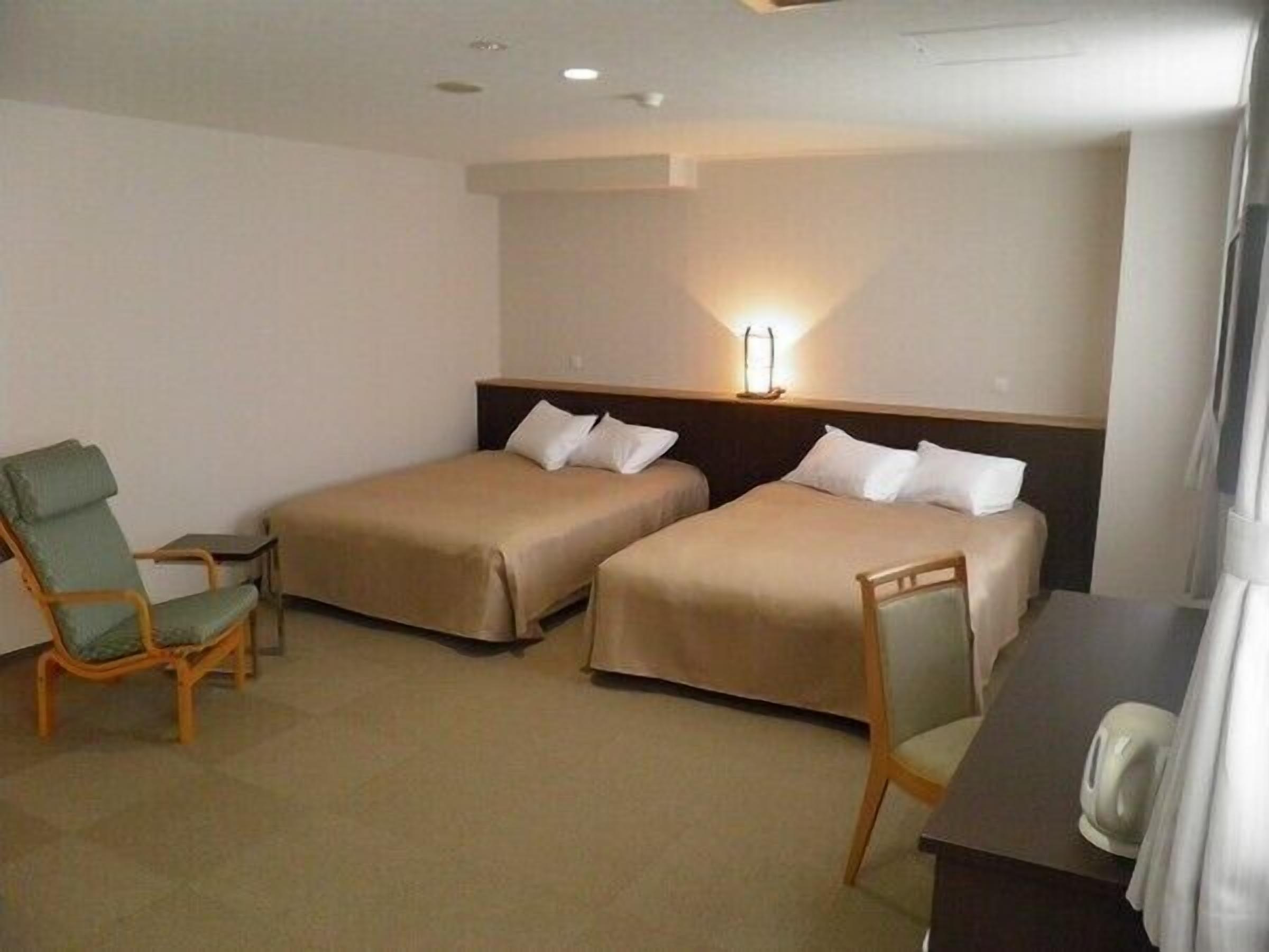 A hotel room with two beds, a desk, a chair, and a lamp.