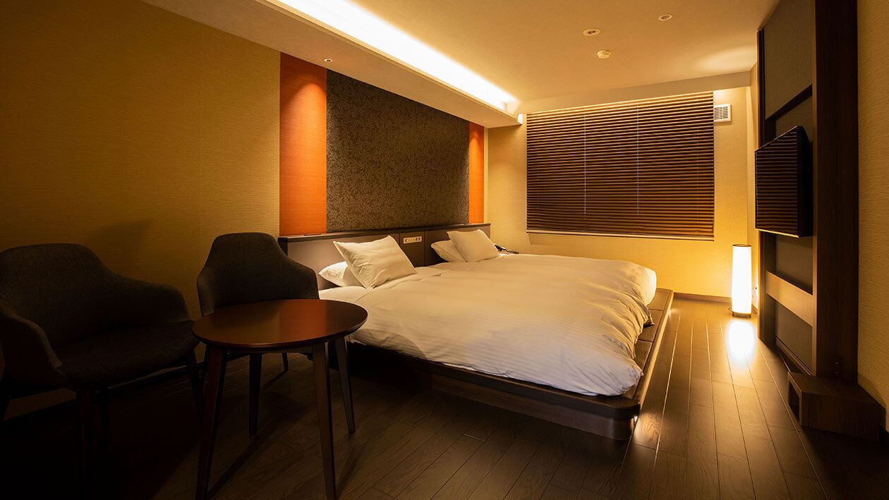 A hotel room with a bed, two armchairs, a small table, a TV, and a window with blinds.