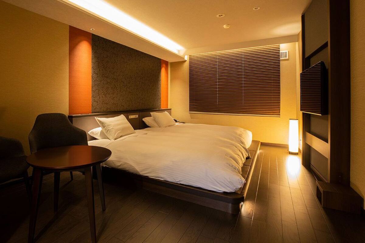 Une chambre d’hôtel comprenant un lit, deux fauteuils, une petite table, un téléviseur et une fenêtre avec des stores.