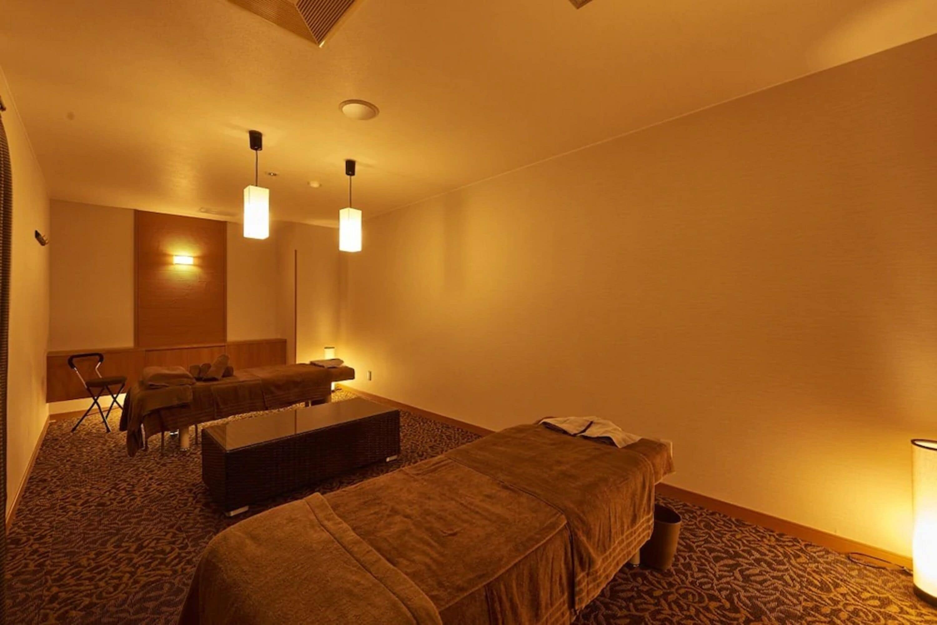 Habitación de hotel con dos camas individuales, una camilla de masaje, un pequeño rincón de descanso y un espejo grande.