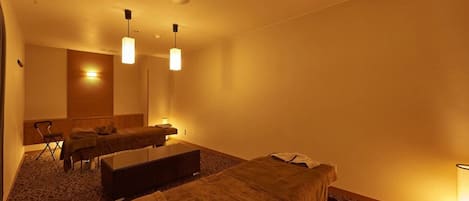 A hotel room with two single beds, a massage table, a small seating area, and a large mirror.