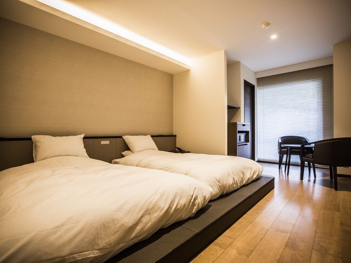 A hotel room with two beds, a desk, a chair, and a large window with blinds.