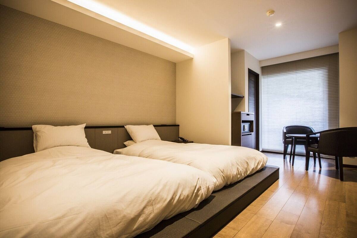 Une chambre d’hôtel avec deux lits, un bureau, une chaise et une grande fenêtre avec des stores.