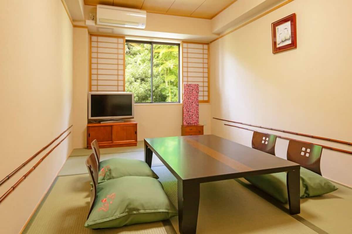 Una habitación japonesa tradicional con suelo de tatami, una mesa baja, cojines, un televisor y una ventana con vistas a la vegetación.