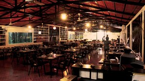 Um restaurante espaçoso com pé direito alto e vigas aparentes, com várias mesas e cadeiras, janelas amplas e vista para o exterior.