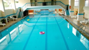 Uma piscina interior ampla, com pistas demarcadas, um dispositivo de flutuação central e várias espreguiçadeiras ao redor.