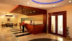 Uma área de recepção de hotel, com balcão, sala de espera com sofá e poltronas, e escadaria para um andar superior.