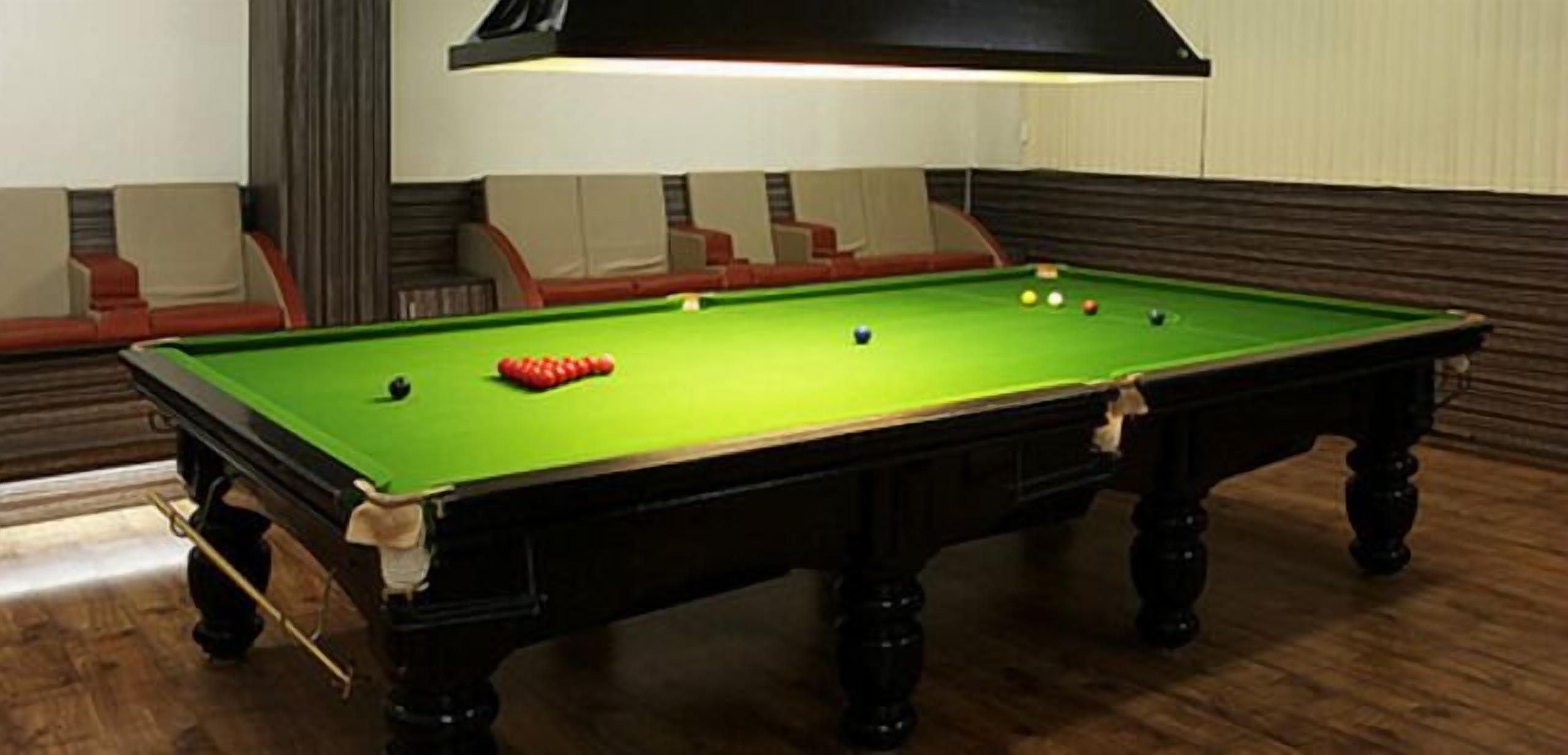 Une table de billard avec des billes, une queue de billard et une bille rouge, dans une pièce au plancher en bois et dotée de sièges rembourrés.