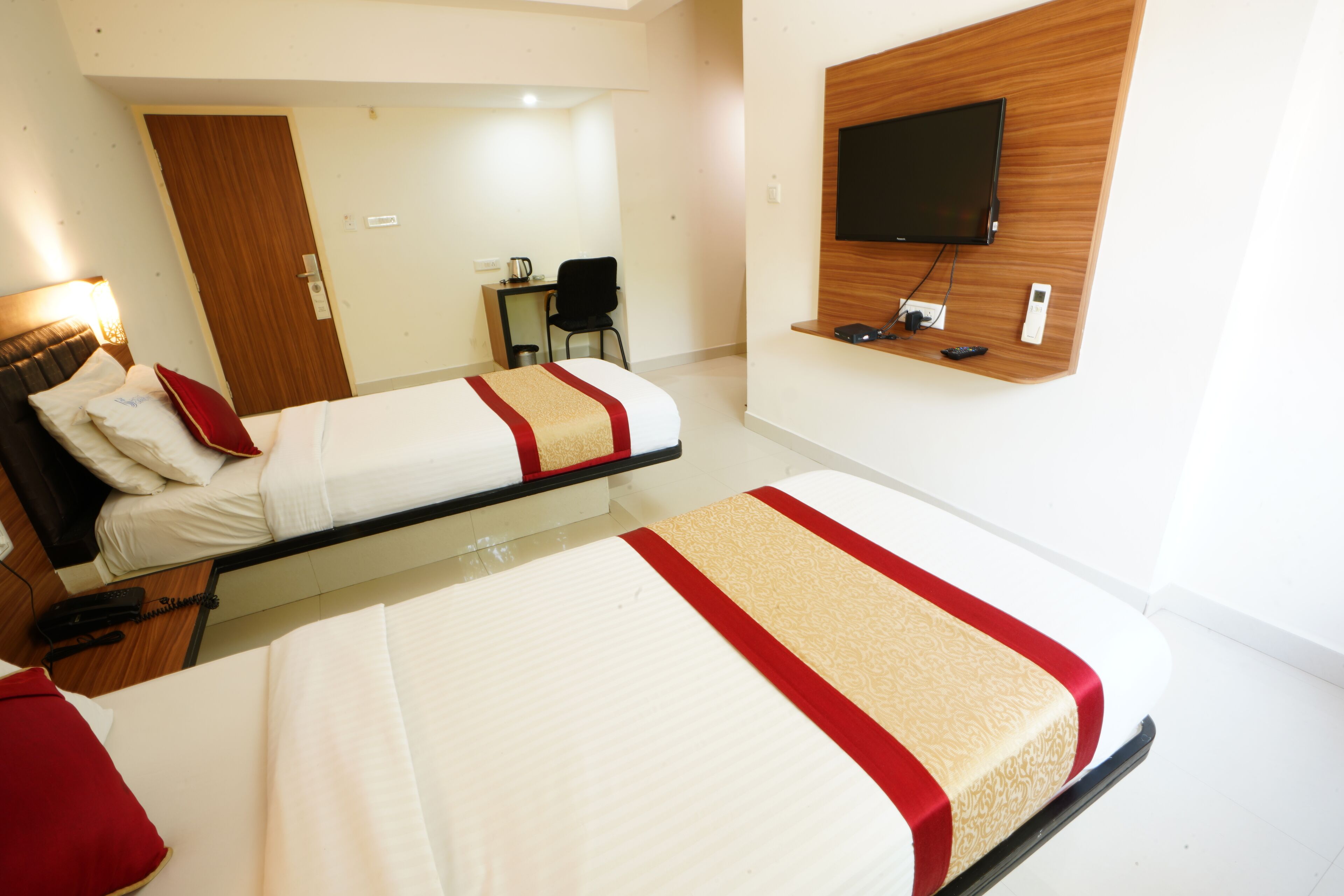 Habitación de hotel con dos camas, un televisor montado en la pared, un escritorio con una silla y una puerta.