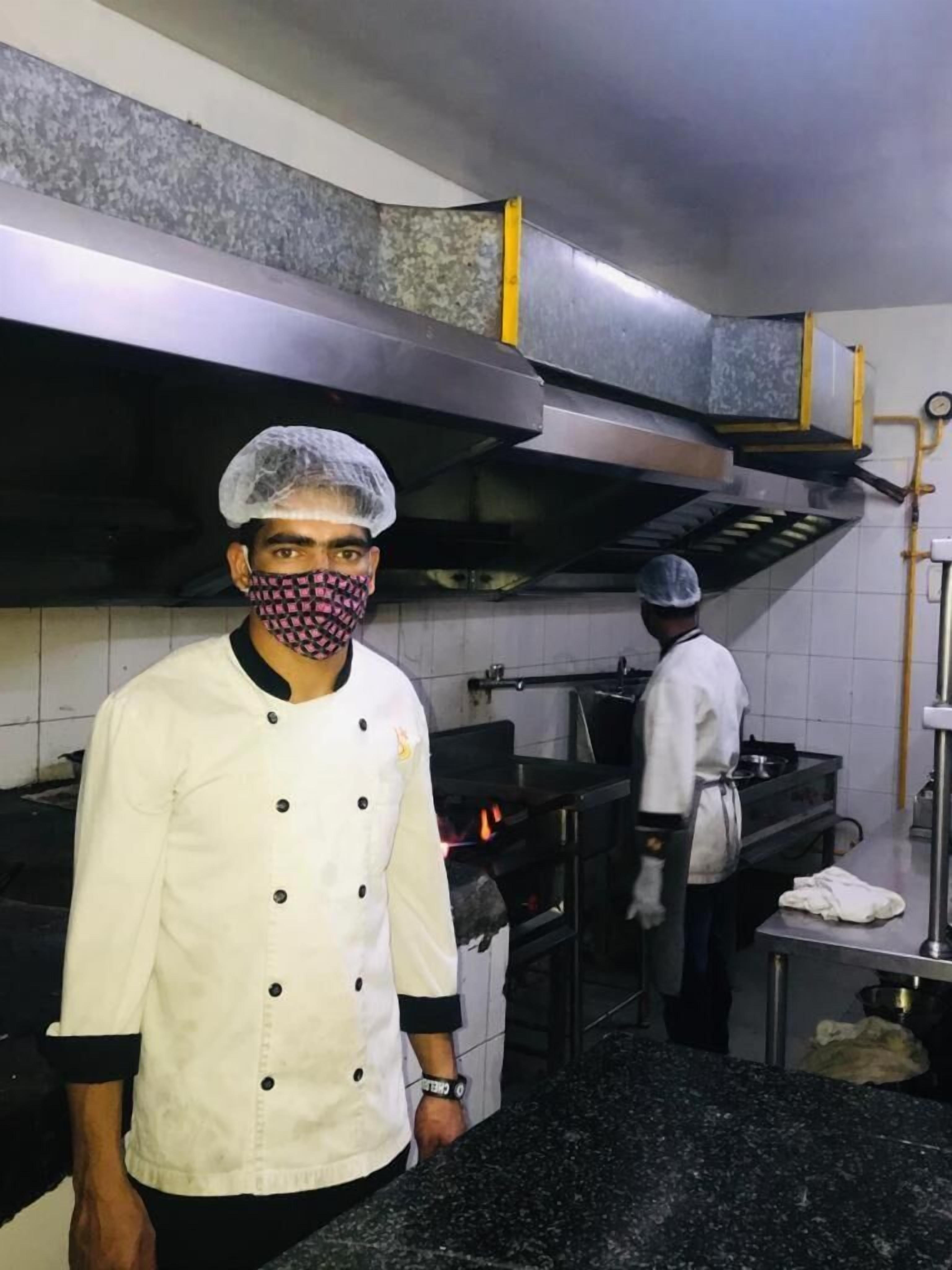 Un chef, vêtu d’un uniforme blanc et coiffé d’un filet à cheveux, se tient dans une cuisine dotée de plans de travail en acier inoxydable et d’une hotte au-dessus de la cuisinière.