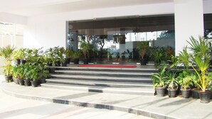 Una entrada moderna de edificio con fachada de vidrio, plantas en macetas y una alfombra roja que conduce a la entrada.