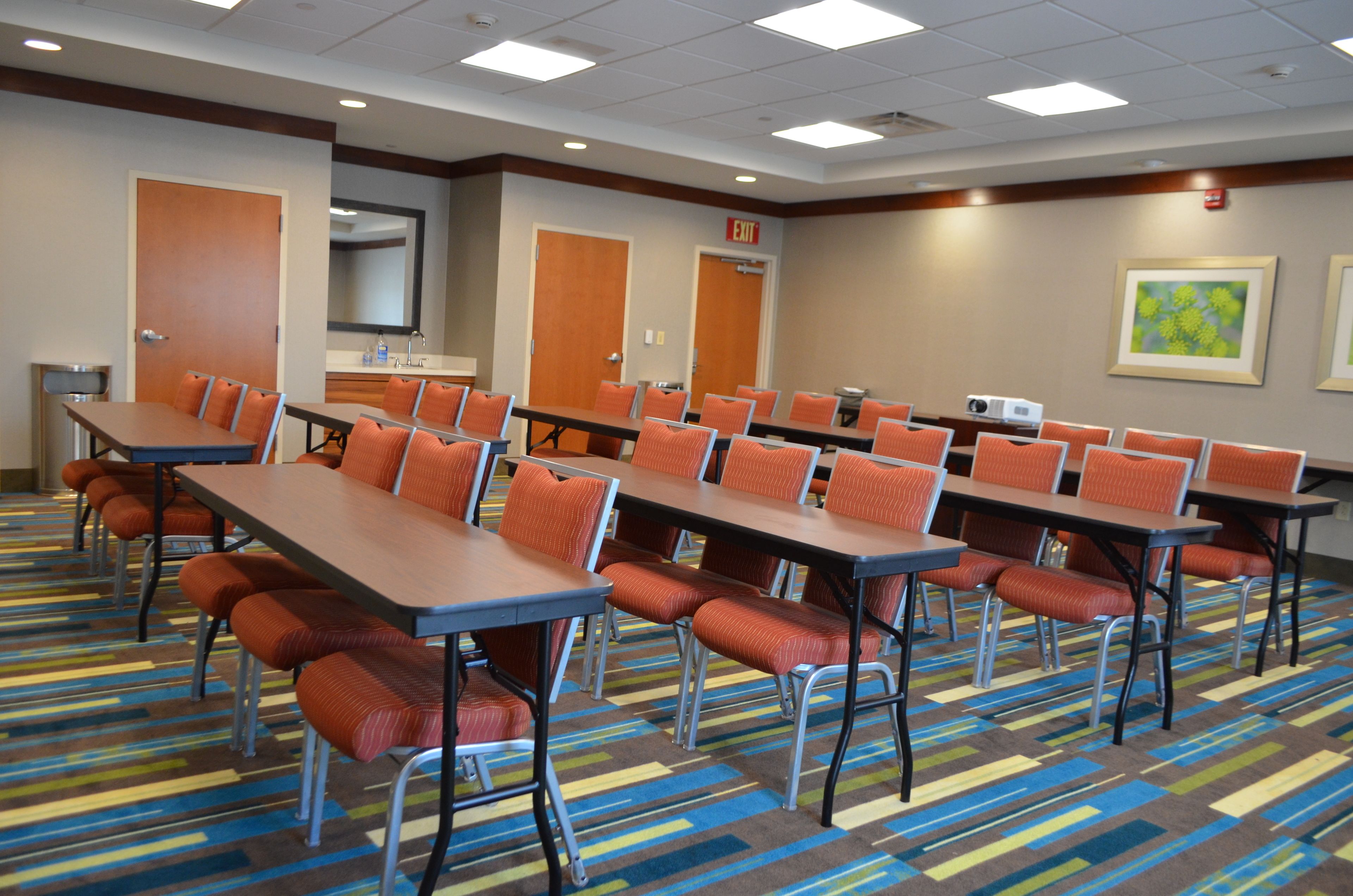 Sala de conferências com várias mesas e cadeiras, equipada com um projetor e um quadro branco.