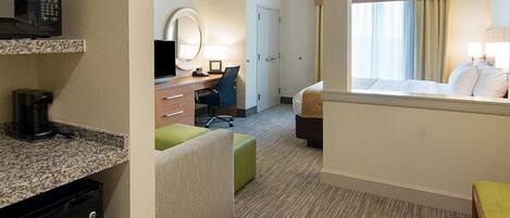 A hotel room with a kitchenette, a desk with a chair, a bed, a window with curtains, and a mirror.