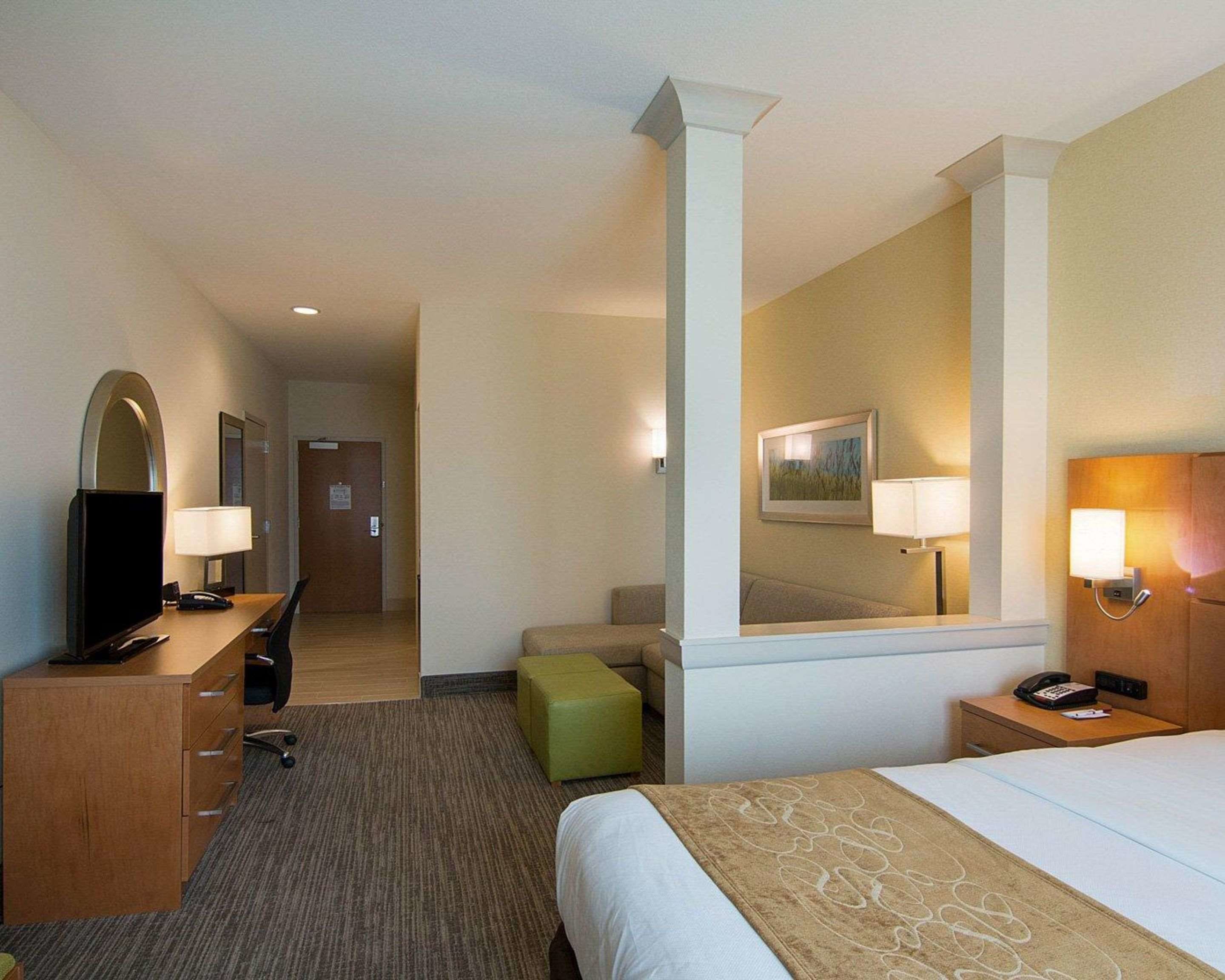 A hotel room with a bed, a desk with a chair, a television, a green ottoman, and a mirror.