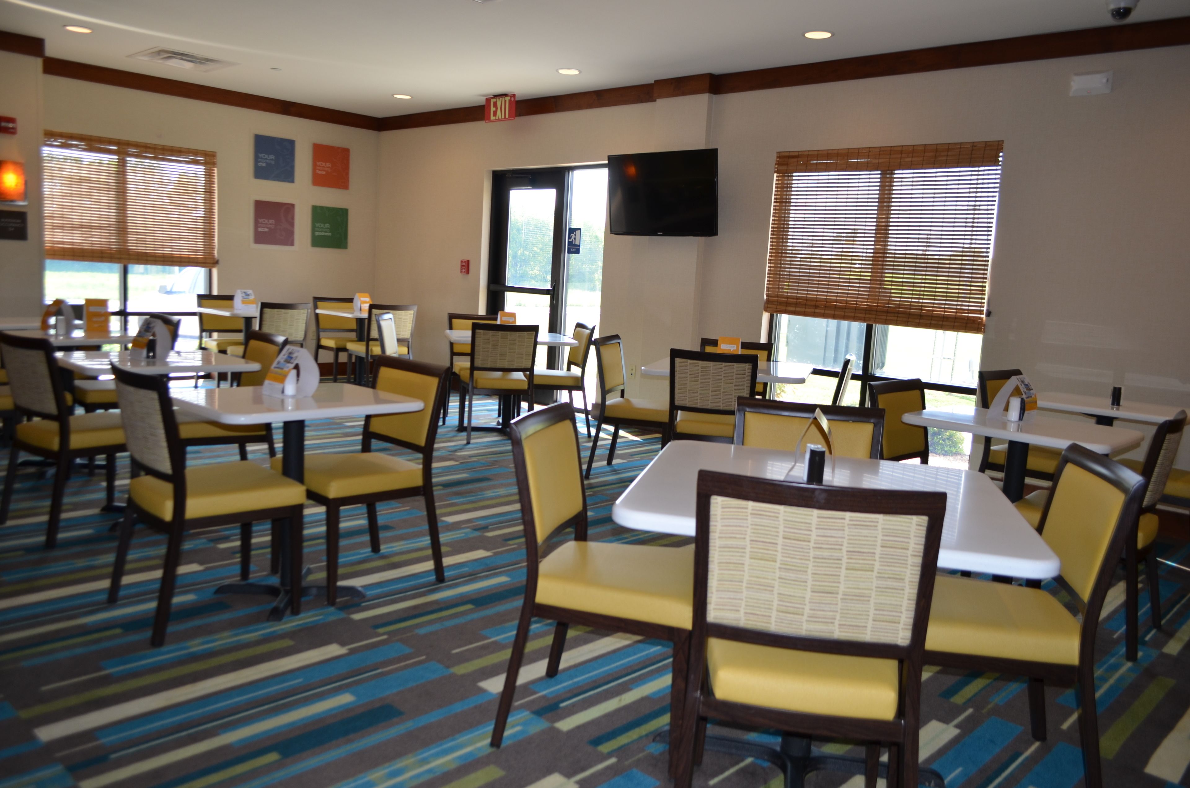 A hotel dining area with multiple tables and chairs, a flat-screen TV mounted on the wall, and large windows with blinds.