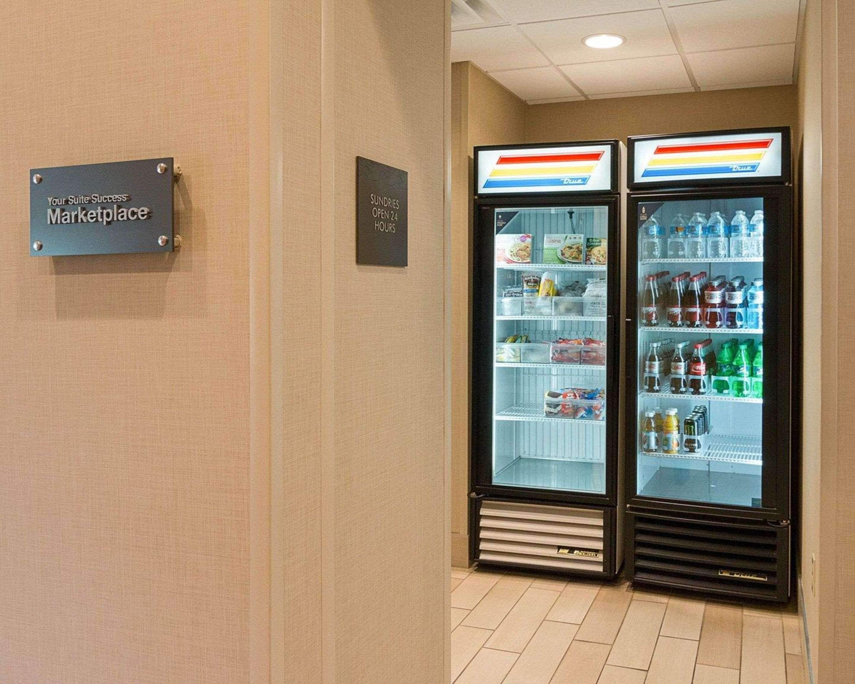 Una zona de kitchenette con un cartel que dice "Your Suite Success Marketplace" y un refrigerador de artículos de conveniencia abierto las 24 horas, lleno de bebidas y aperitivos.