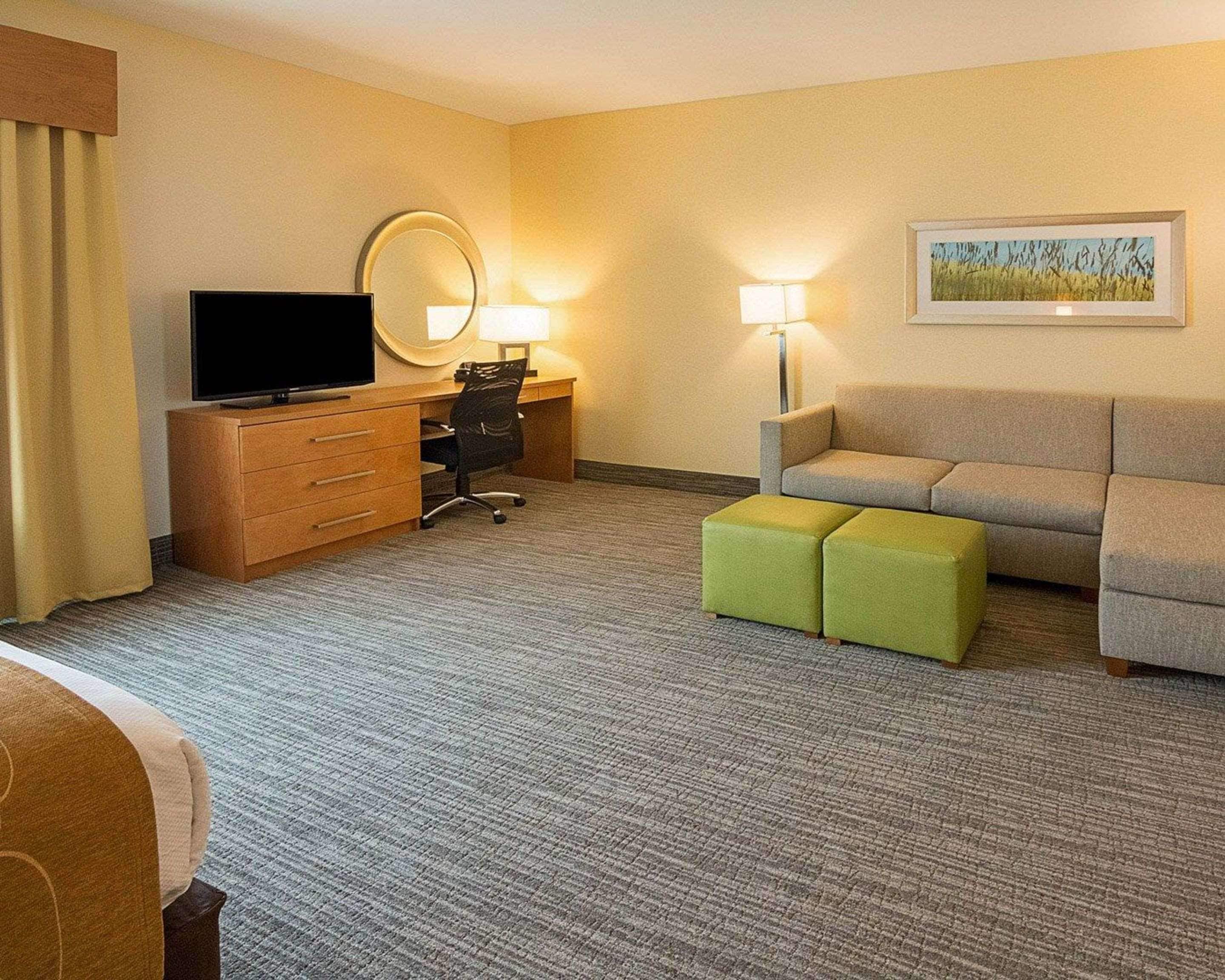 A hotel room with a flat-screen TV, a desk with a chair, a sofa, two ottomans, a lamp, and a painting on the wall.