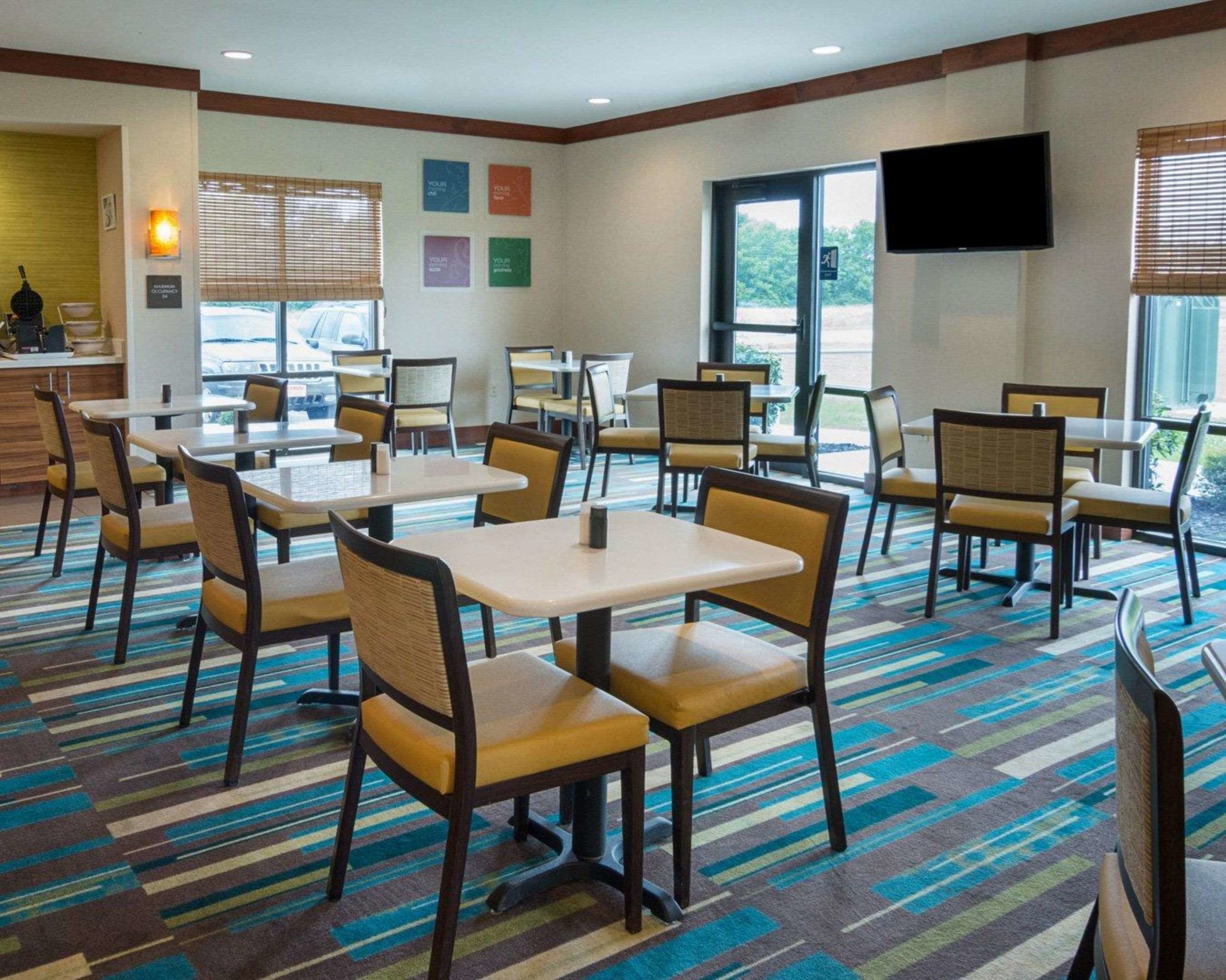 Een restaurantgedeelte in een hotel met meerdere tafels en stoelen, een aan de muur bevestigde flatscreen-tv, en een raam met uitzicht op de omgeving.
