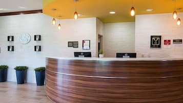 A reception area with a curved wooden reception desk, a white brick wall, and a yellow ceiling. There are several wall-mounted light fixtures and a few potted plants.