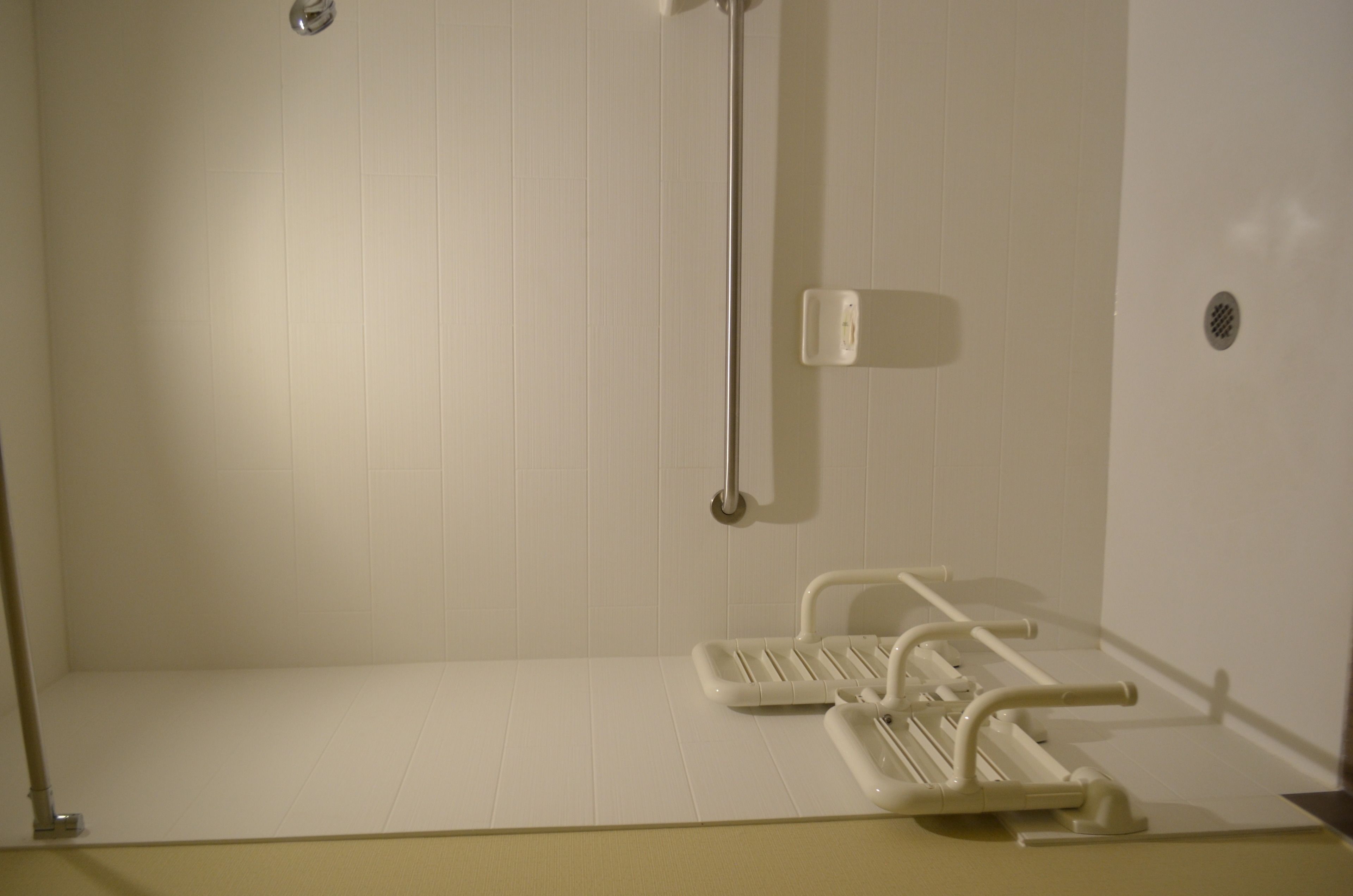 Un baño con azulejos blancos, ducha con cabezal de ducha manual y inodoro con barra de apoyo.