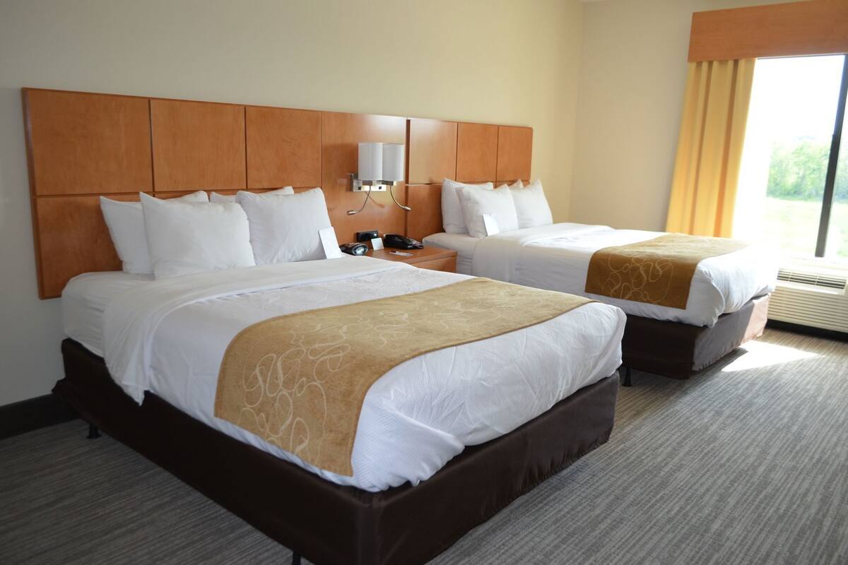 A hotel room with two beds, a wooden headboard, a bedside table with a lamp, and a window with curtains.
